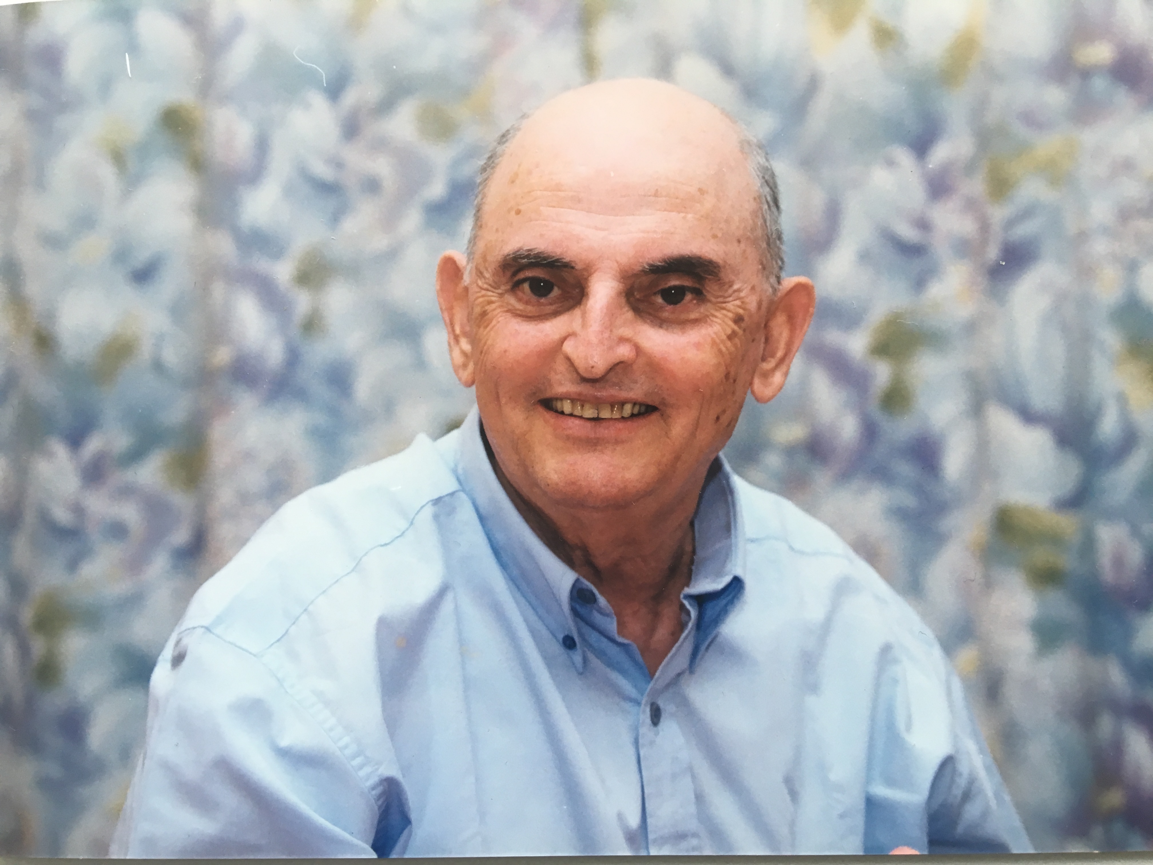 Photograph of the artist Asher Kalderon: an older, smiling bald man in a pale blue shirt, seated before a soft floral-patterned backdrop.